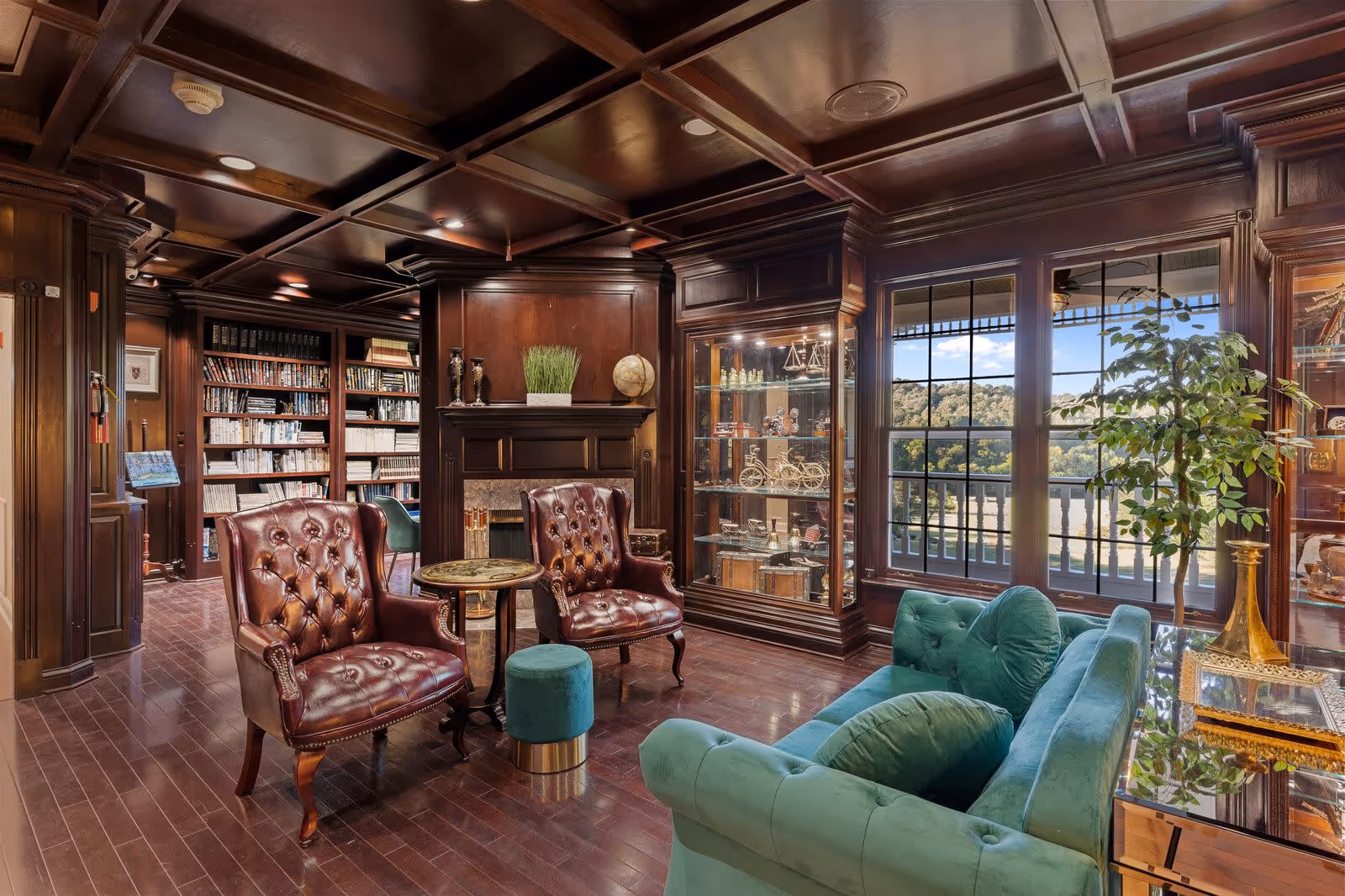A cozy senior living facility lounge with dark wood paneling and ceiling beams, featuring two brown leather armchairs, a small round table, a green velvet sofa with matching cushions, a glass display cabinet with decorative items, a fireplace, a bookshelf filled with books, and large windows showing a scenic outdoor view with trees and hills.