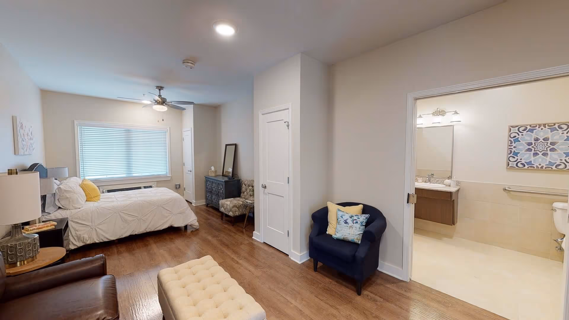 A senior living facility bedroom with a single bed dressed in white bedding and a yellow pillow, a nightstand with a lamp, a ceiling fan, a window with blinds, a blue armchair with two pillows, a patterned chair, a dresser with a mirror, and an open door leading to a bathroom with a sink, toilet, and decorative wall art.