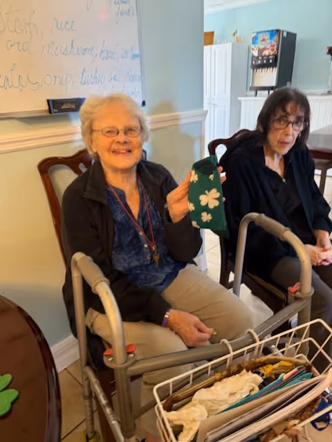 Two elderly women sitting in chairs in a room. One woman is smiling and holding a green item with white shamrocks. A walker is in front of her. Behind them is a whiteboard with writing and a beverage dispenser.