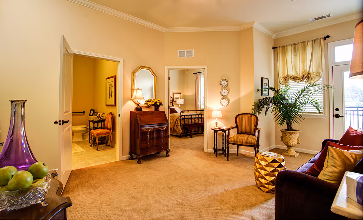 A cozy living area in a senior living facility featuring a purple sofa with colorful pillows, a gold geometric side table, a potted plant near a window with sheer curtains, and a small wooden table with a lamp and a striped armchair. Two open doorways reveal a bathroom with a toilet and a bedroom with a bed and nightstand. A wooden chest with a mirror and lamp is positioned between the doorways.