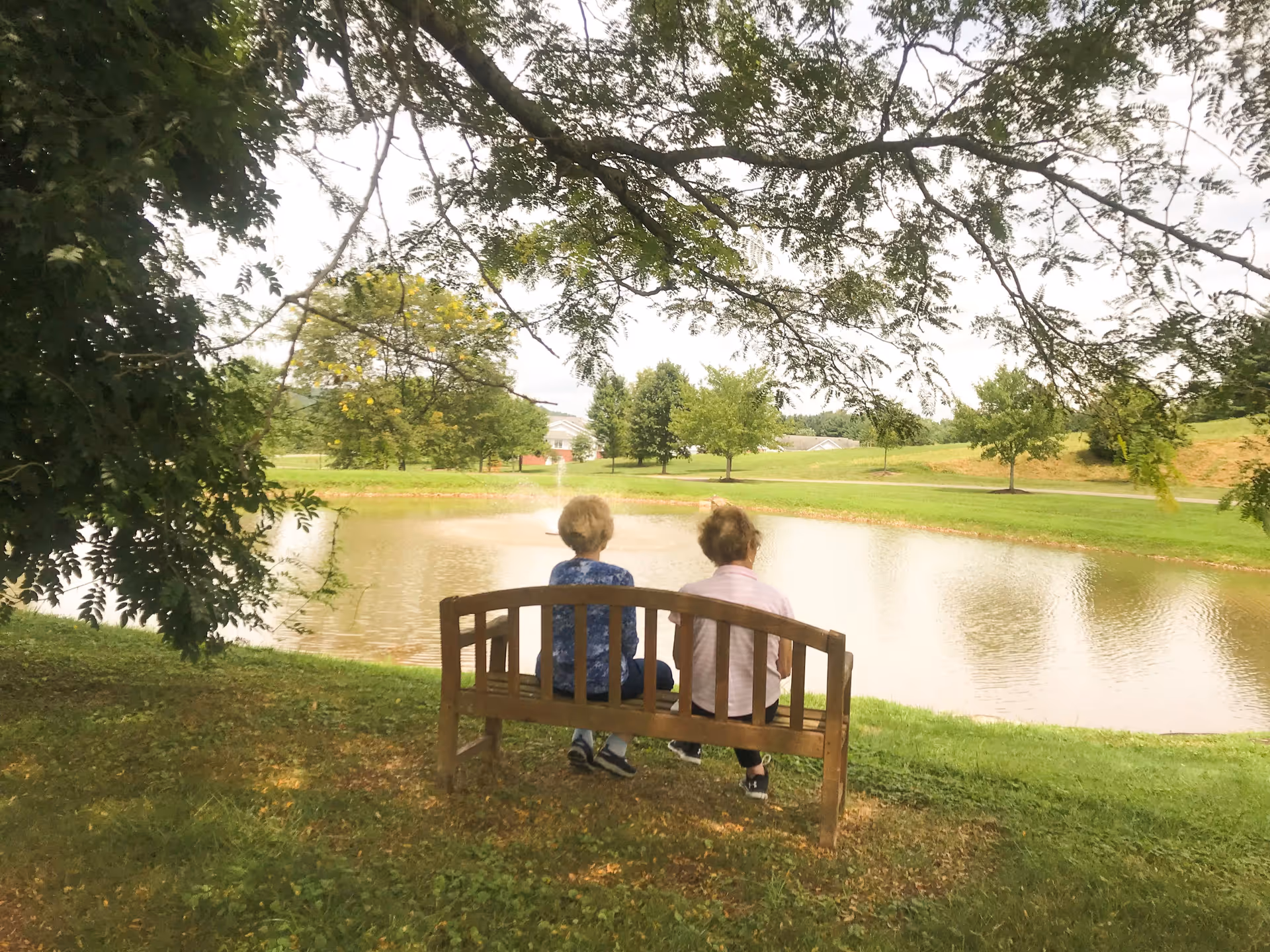 Two elderly women sitting on a wooden bench under a tree, facing a small pond with a fountain in a green park-like setting.