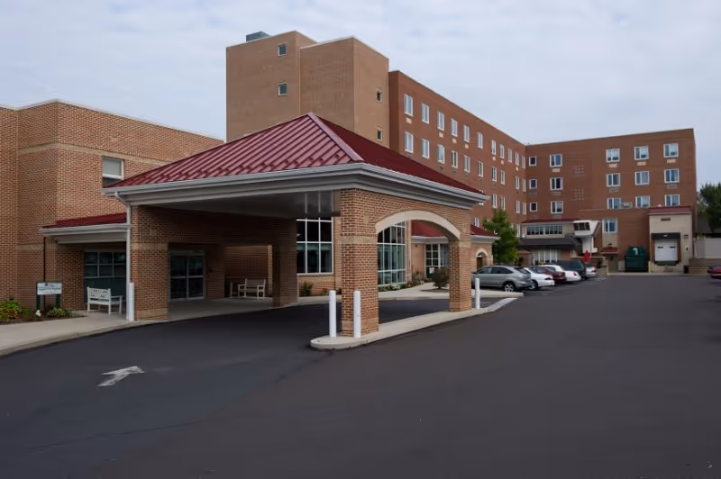 Exterior view of a multi-story brick senior living facility with a covered entrance and several parked cars in the parking lot under an overcast sky.