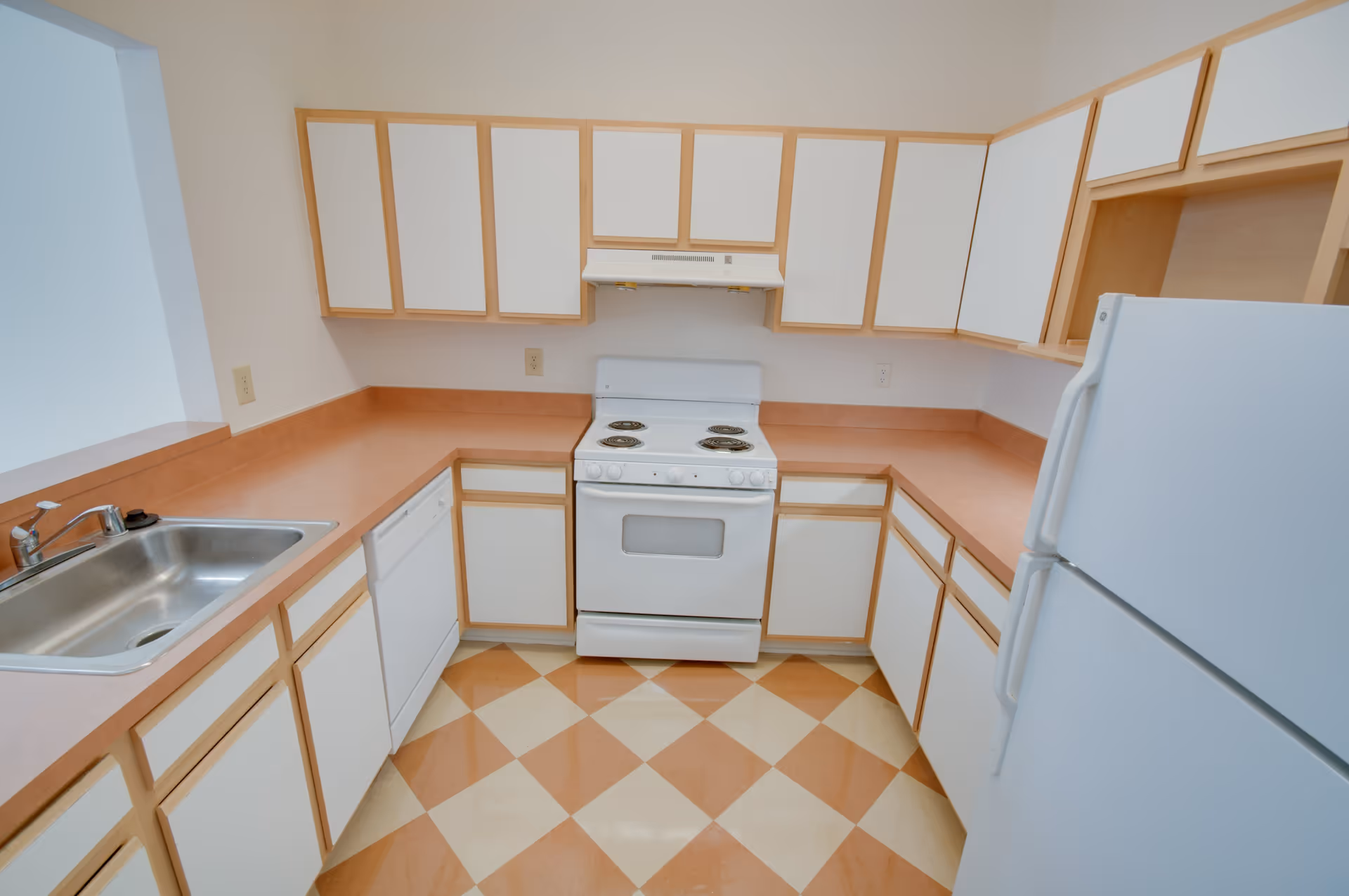 A compact kitchen with white cabinets and light wood trim, featuring a white electric stove with four burners, a white refrigerator, a dishwasher, a stainless steel sink, and peach-colored countertops. The floor has a checkered pattern with peach and cream tiles.