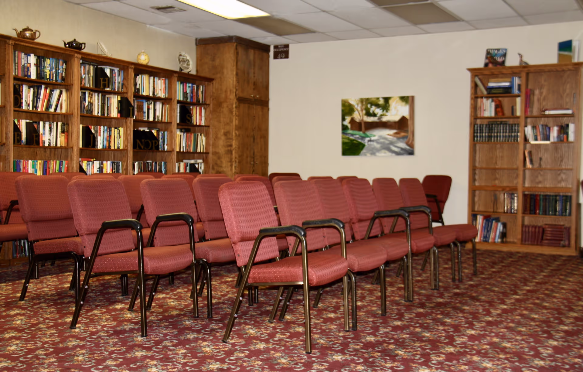 Room with rows of red cushioned chairs arranged facing forward, surrounded by wooden bookshelves filled with books. The room has a patterned carpet and a painting on the wall.