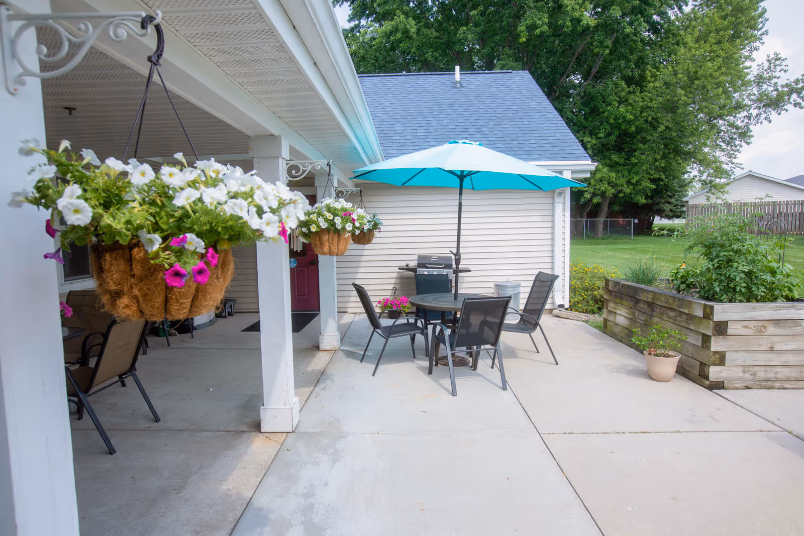 Outdoor patio with hanging flower baskets, a round table and aqua umbrella surrounded by chairs, a grill, and raised planters.