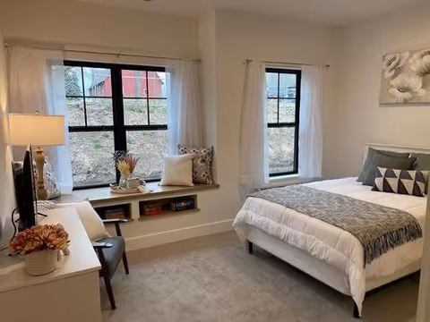 A cozy bedroom with a neatly made bed featuring white and gray bedding, a window seat with cushions and books underneath, a small desk with a chair, a table lamp, and decorative flowers. Two windows with white curtains let in natural light.
