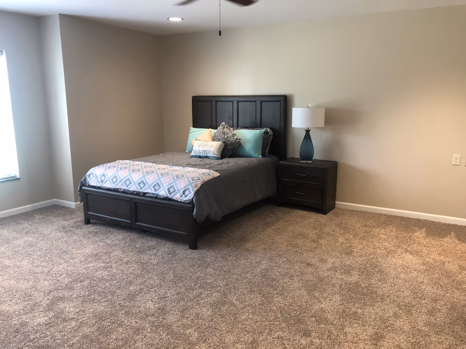 A bedroom with a dark wooden bed frame and headboard, a bed made with gray bedding, decorative pillows, and a patterned throw blanket. Next to the bed is a matching dark wooden nightstand with a blue lamp. The room has beige walls, a window on the left side, and carpeted flooring.