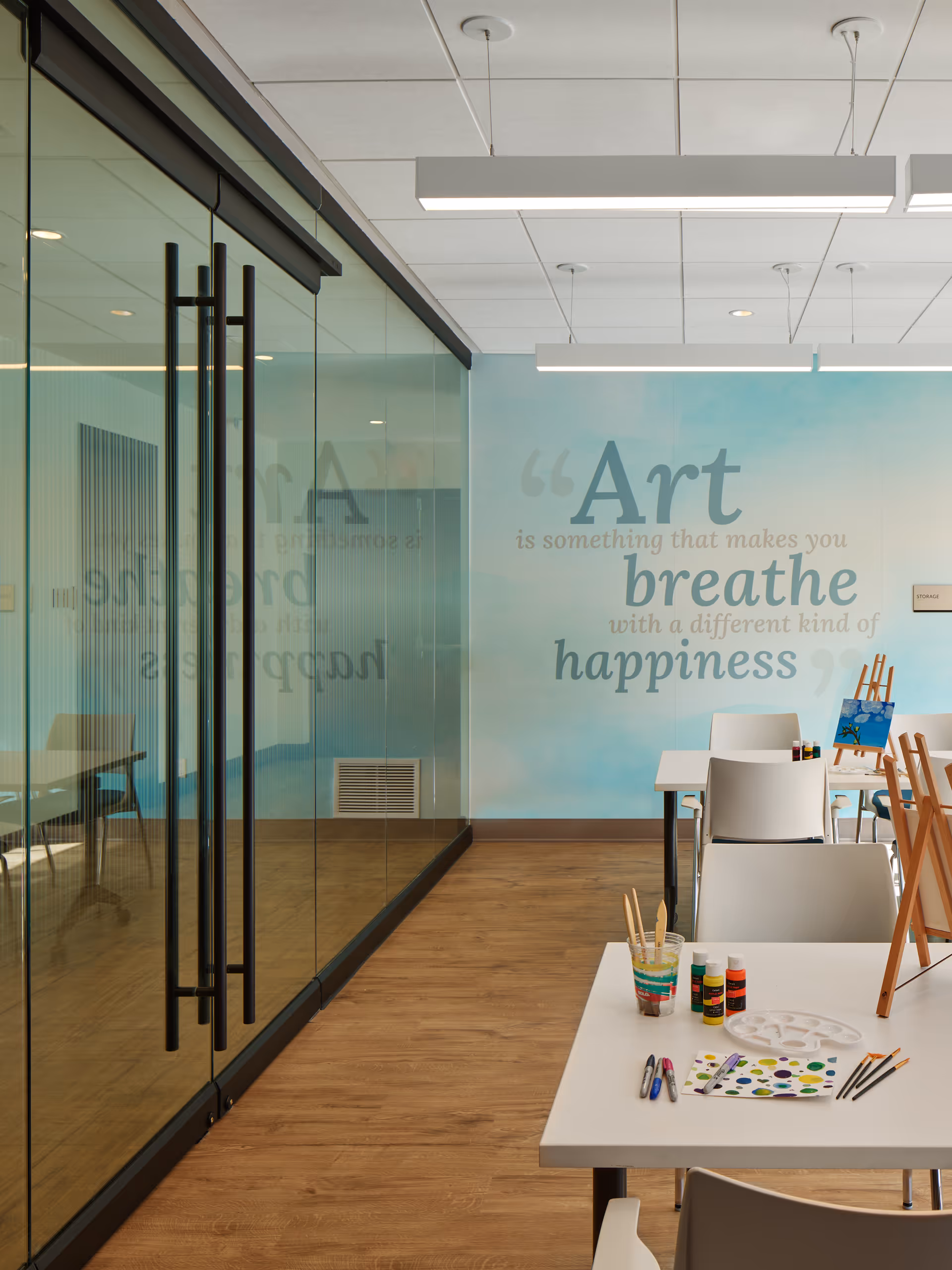 Interior view of a bright art room with wooden flooring, white tables and chairs, painting supplies including brushes, paint bottles, and a palette on the tables. A large glass wall with black handles is on the left side. The far wall has a blue gradient background with a quote that reads, 'Art is something that makes you breathe with a different kind of happiness.'