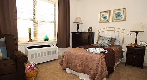 A neatly furnished bedroom with a single bed, bedside tables and lamps, a dresser, armchair, window with curtains, and framed floral artwork.