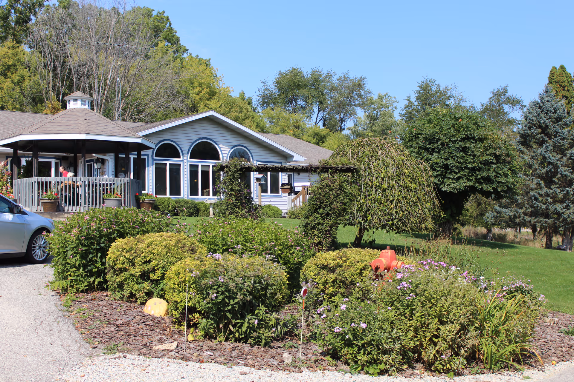 Exterior view of a single-story building with large arched windows and a gazebo on the left side. The foreground features a garden with various bushes, flowers, and a red fire hydrant. Trees and a clear blue sky are visible in the background.