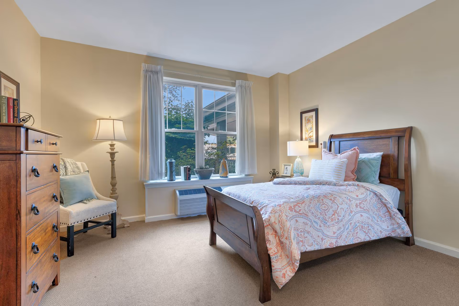 Bright furnished bedroom featuring a wooden bed with patterned bedding, a dresser, armchair, lamps, and a window with curtains.