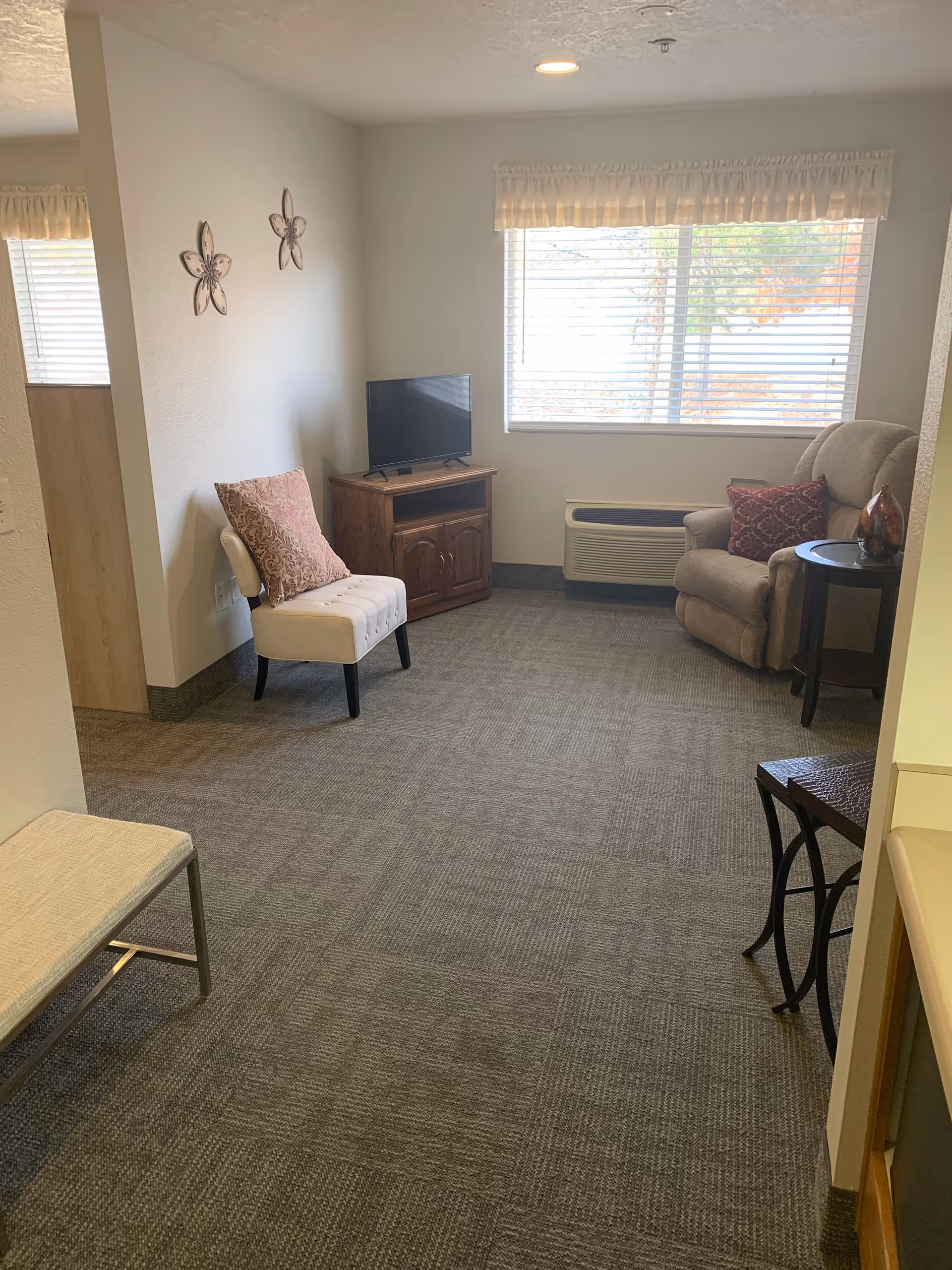 A small living room area with a beige recliner chair with a red patterned pillow, a white cushioned chair with a pink pillow, a wooden TV stand with a flat-screen TV, a small round side table with a decorative item, and a large window with blinds and a valance. The room has gray carpet and light-colored walls with two flower-shaped wall decorations.