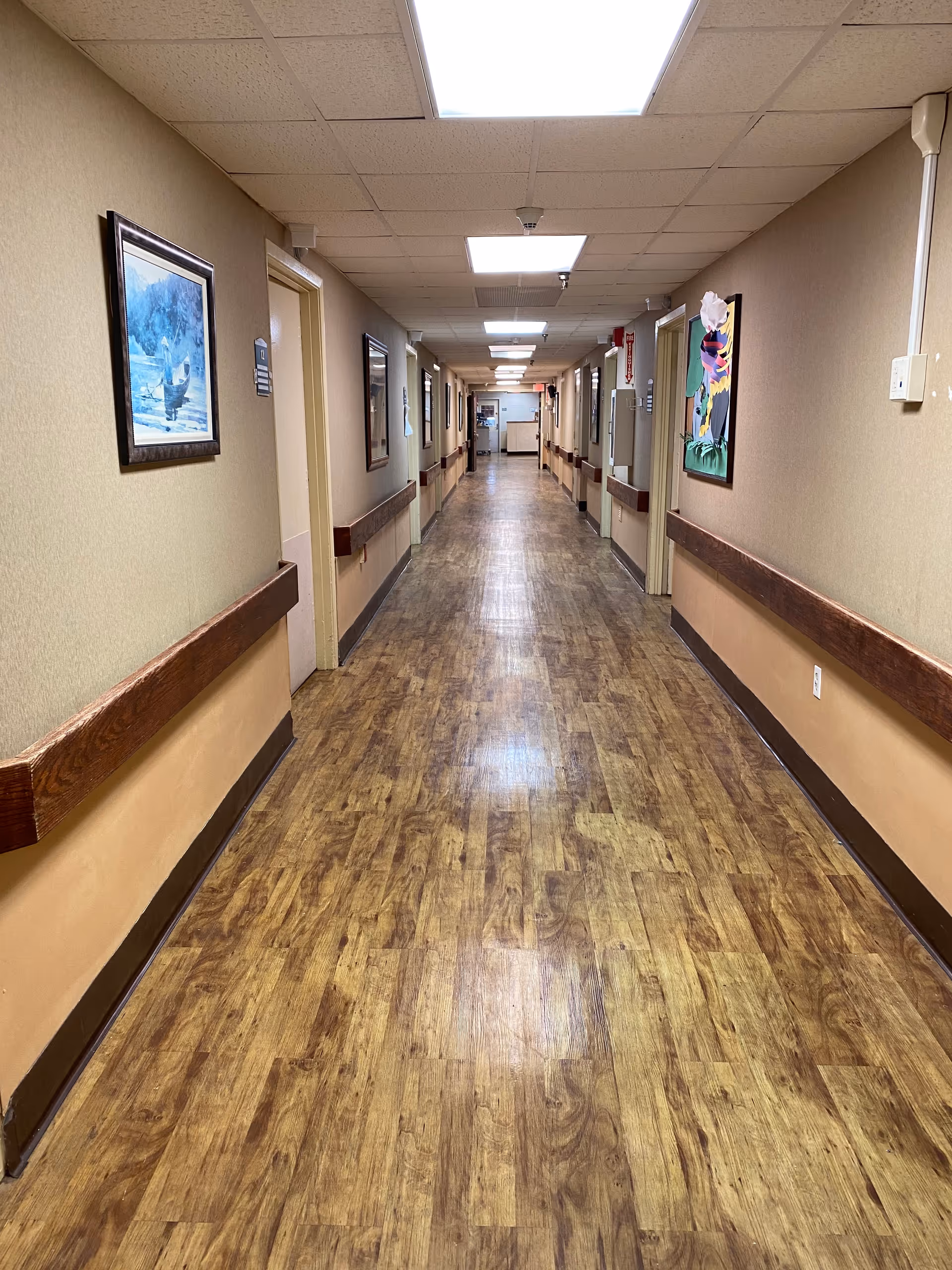 A long, well-lit hallway in a healthcare facility with wood-patterned flooring, beige walls, handrails on both sides, and several framed paintings hanging on the walls. Multiple doorways line the corridor, and ceiling lights illuminate the space.