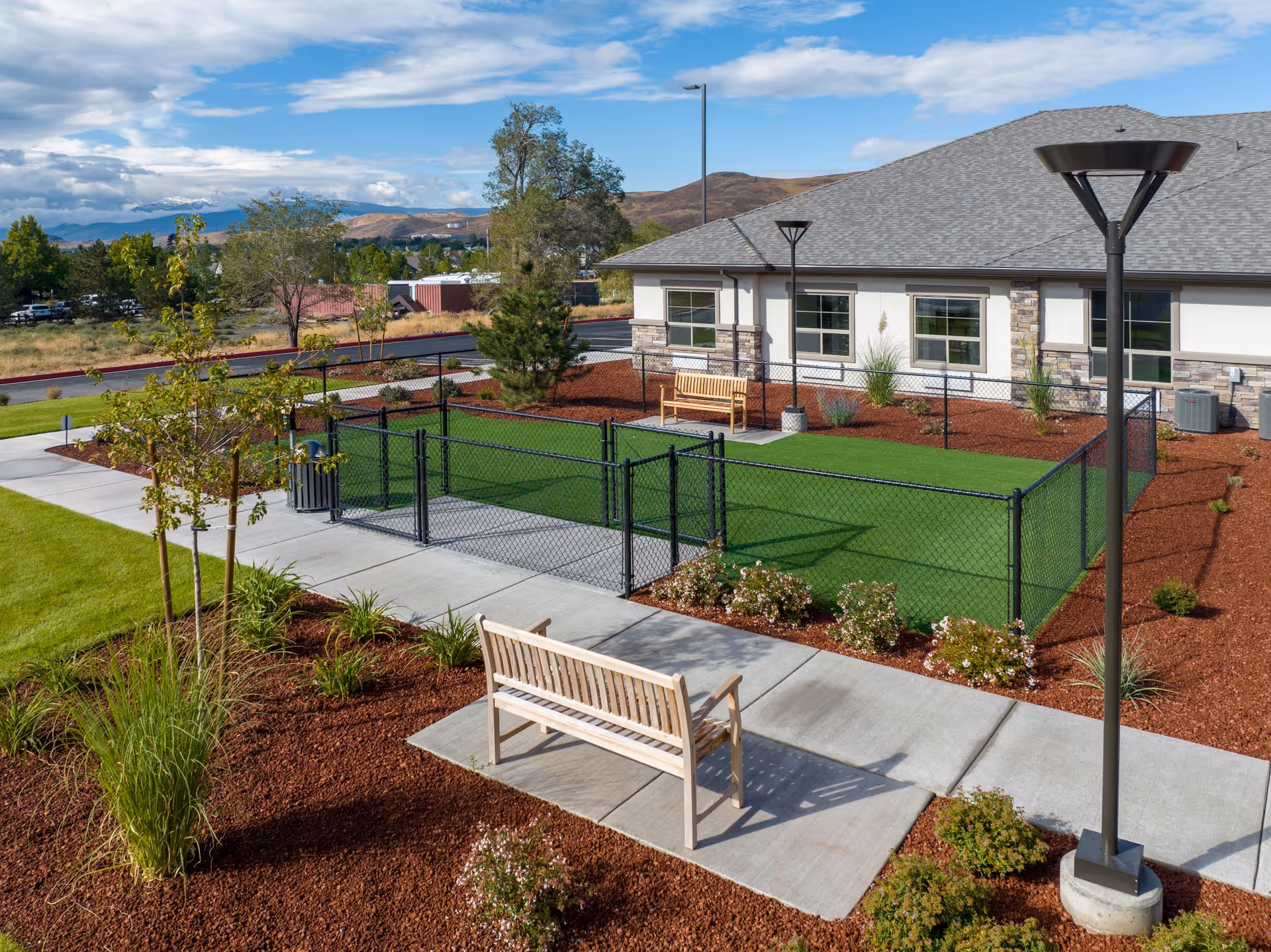 Outdoor area of Kiley Ranch Senior Living featuring a fenced green lawn, two wooden benches on concrete pads, landscaped garden beds with mulch and plants, a paved walkway, and a building with stone and stucco exterior under a partly cloudy sky.