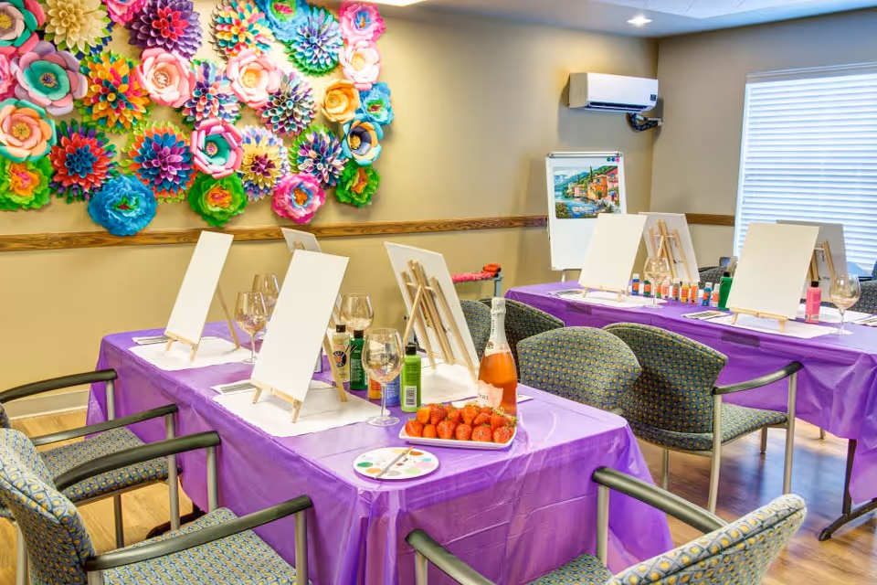 A bright and colorful art activity room with tables covered in purple tablecloths, each set with blank canvases on easels, paint bottles, wine glasses, and a plate of strawberries. The wall is decorated with large, vibrant paper flowers, and there is a window with blinds letting in natural light.
