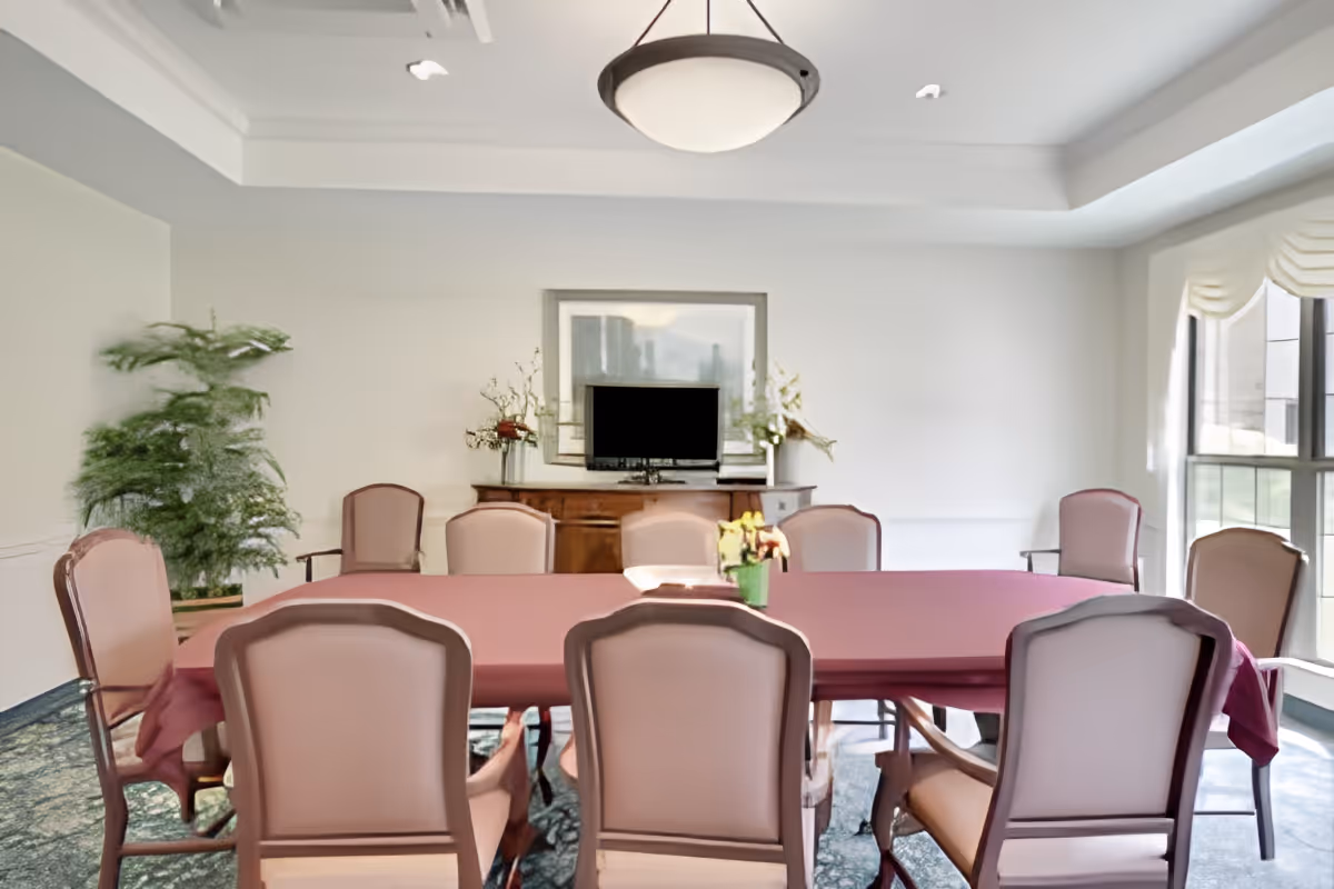 A well-lit dining room with a large rectangular table covered with a maroon tablecloth, surrounded by ten upholstered chairs. There is a wooden sideboard against the back wall with a flat-screen TV and decorative plants on it. A large window on the right side lets in natural light, and a potted plant is placed in the left corner of the room. The ceiling features a modern light fixture.