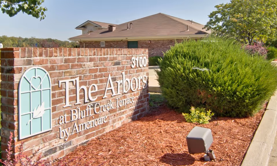 Brick sign for The Arbors at Bluff Creek Terrace by Americare with the address number 3100, surrounded by landscaped bushes and mulch, with a building and trees in the background under a clear sky.