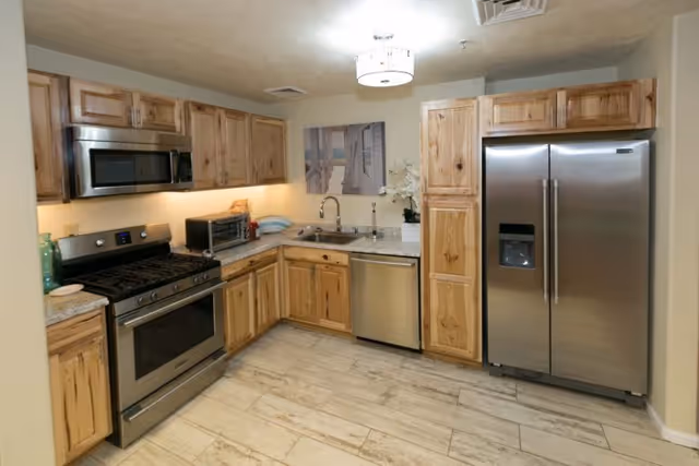 A modern kitchen with wooden cabinets, stainless steel appliances including a refrigerator, oven, microwave, and dishwasher. The kitchen has a light-colored tile floor and a ceiling light fixture. There is a sink with a faucet and a decorative plant on the countertop.