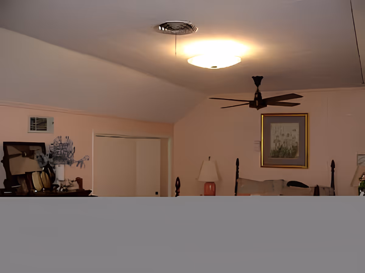 Interior view of a room with a ceiling light and a ceiling fan. The room has light-colored walls with a framed picture hanging on one wall. There is a table lamp on a side table and a dresser with various items on it, including a mirror and decorative objects. A sliding door closet is visible in the background.