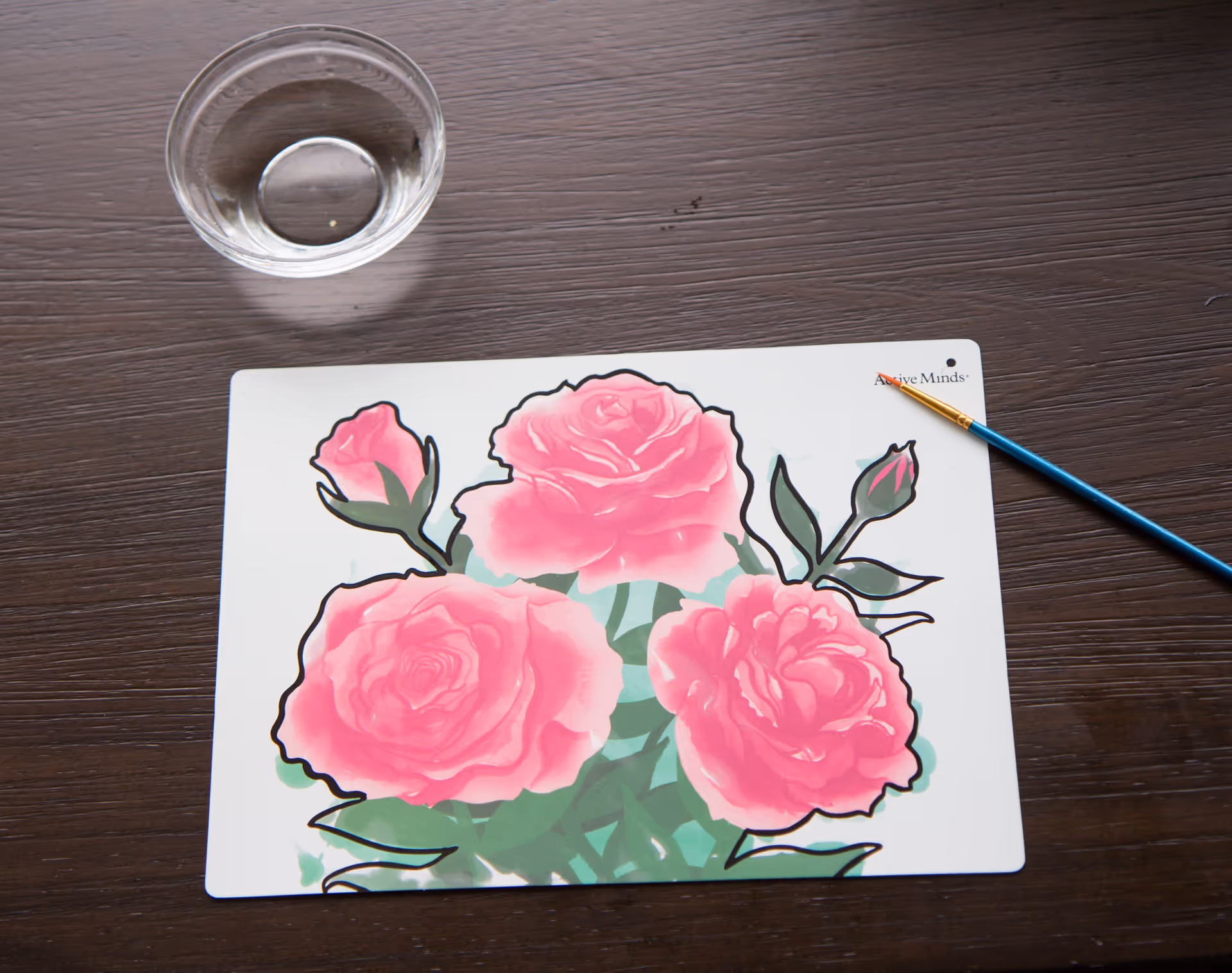 Top-down view of a floral placemat with pink roses on a dark wooden table, with a paintbrush and a small glass bowl.