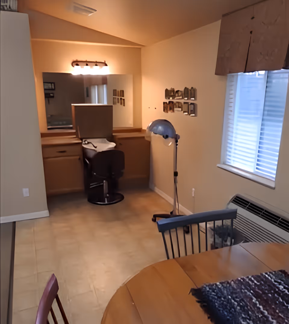 Interior room with a wooden dining table and chairs in the foreground, a window with blinds and a valance on the right, and a vanity area with a mirror, lights, and a salon chair in the background. A standing hair dryer is positioned near the vanity.