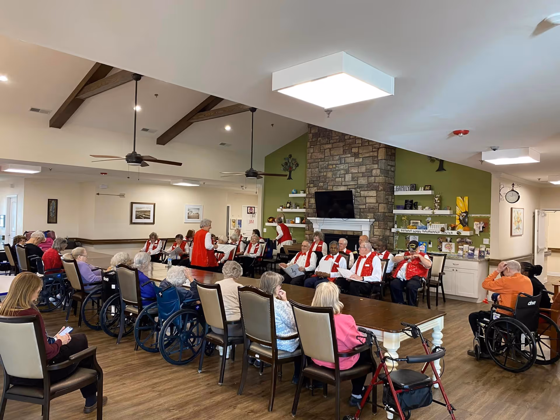 A senior living common room where elderly residents sit in chairs and wheelchairs around tables while a group in red vests performs near a stone fireplace and TV.