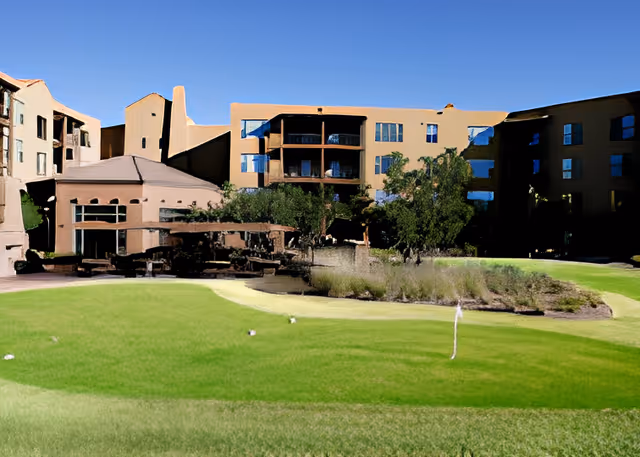 Exterior view of Splendido At Rancho Vistoso senior living facility showing a multi-story building with balconies, surrounded by green lawns, trees, and landscaped bushes under a clear blue sky.