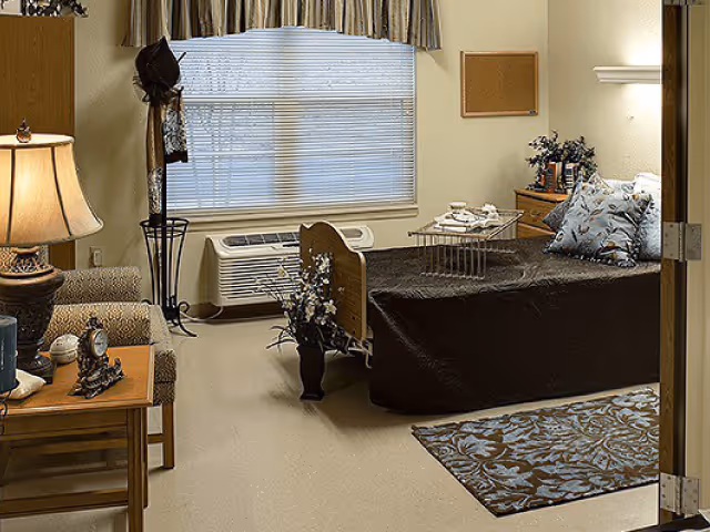 A cozy bedroom in a senior living facility with a single bed covered in a dark bedspread and floral pillows. There is a small wooden nightstand with a lamp, clock, and decorative items. A window with blinds and a valance is behind the bed, and a patterned rug lies on the floor. A chair and a coat rack are also visible in the room.