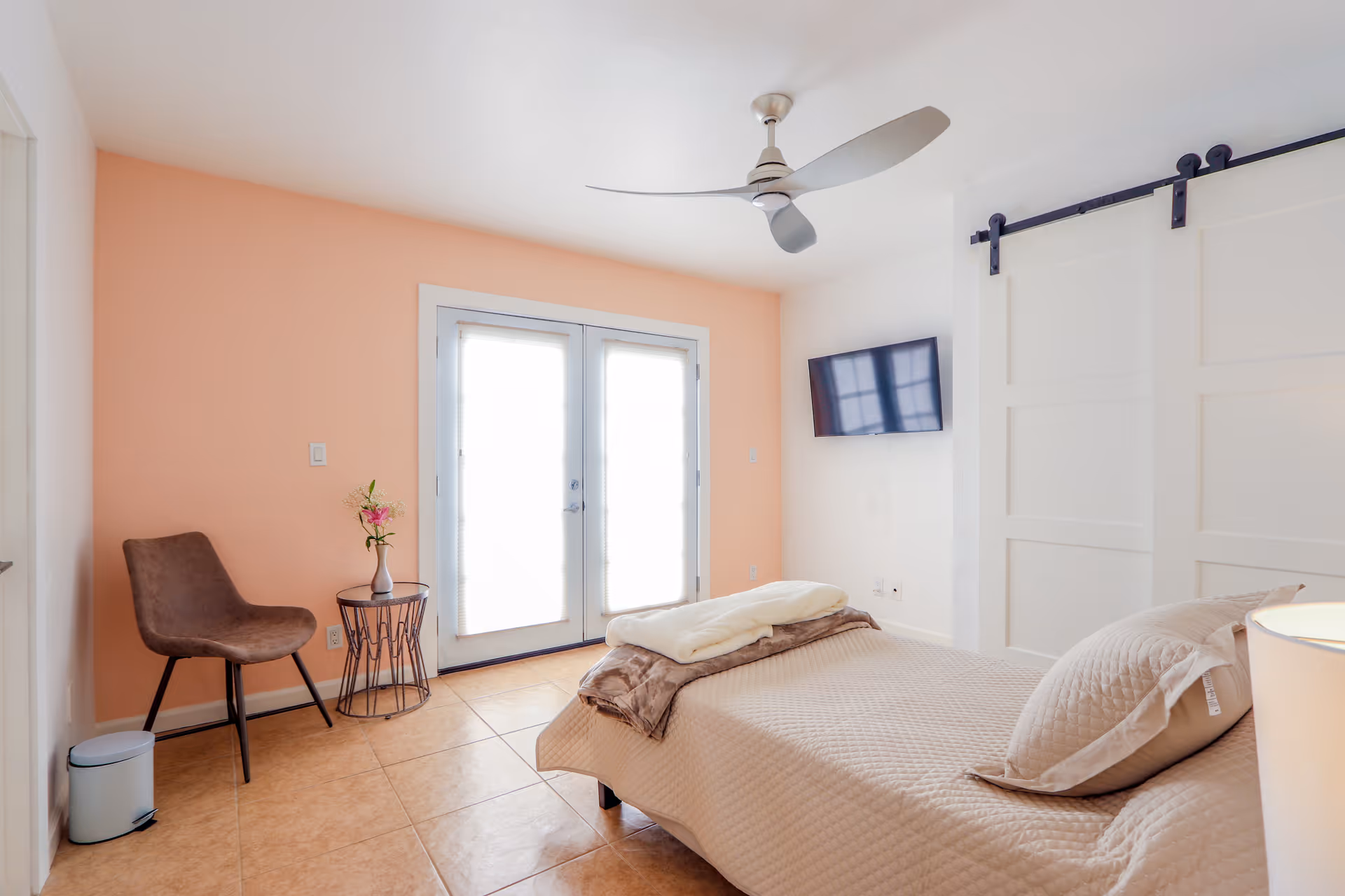 A bright bedroom with a peach accent wall, double glass doors letting in natural light, a bed with beige bedding, a wall-mounted TV, a modern ceiling fan, a brown chair next to a small round table with a vase of flowers, and a sliding barn door.