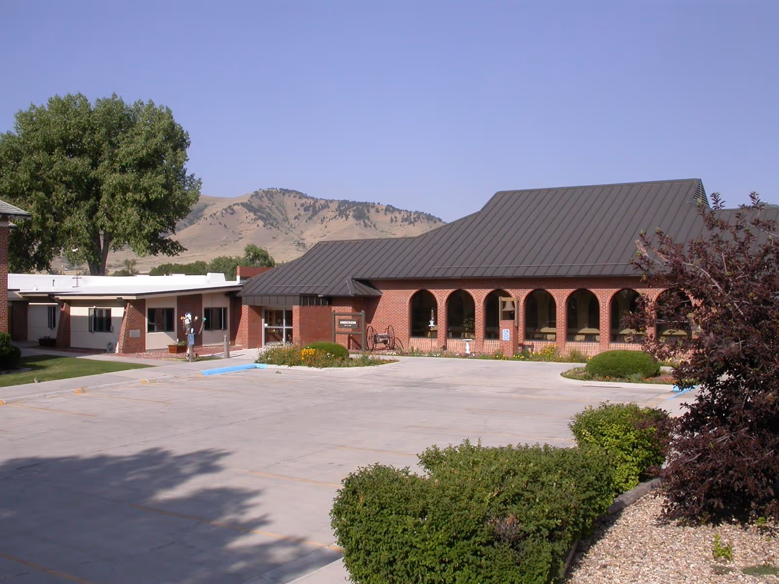 Exterior view of the Veterans' Home of Wyoming building with a brick facade, arched windows, a dark metal roof, and a parking lot in front. There are bushes and trees around the building, with hills visible in the background under a clear blue sky.