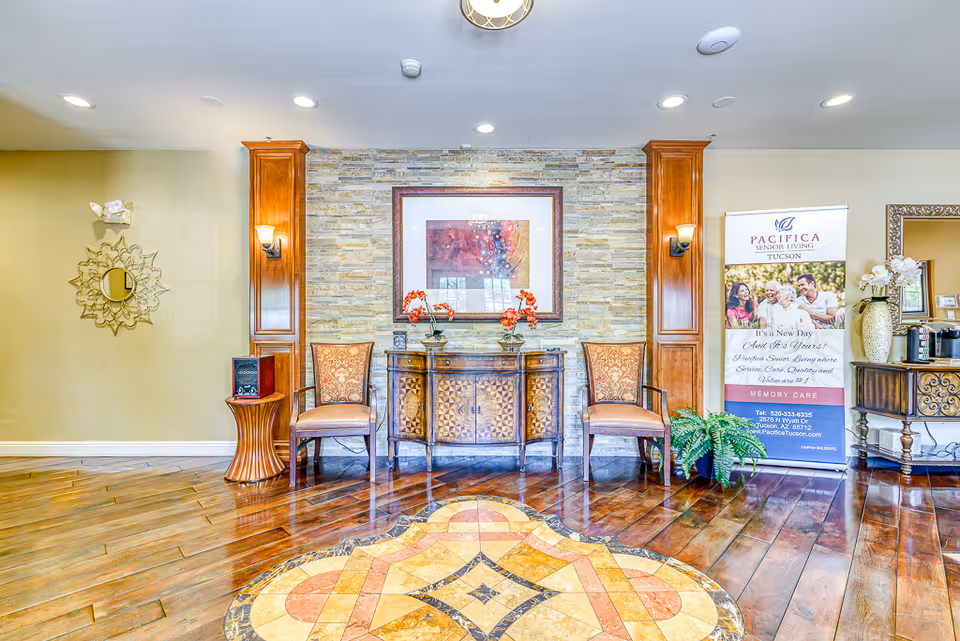 A bright and welcoming interior space with polished wooden floors and a decorative tile inlay. The back wall features a stone accent with a framed abstract painting centered above a wooden cabinet. Two upholstered chairs flank the cabinet, and there are wooden columns with wall sconces on either side. To the right, there is a banner for Pacifica Senior Living Tucson Memory Care, and a small table with a vase of flowers and a coffee machine. A decorative mirror is on the left wall.