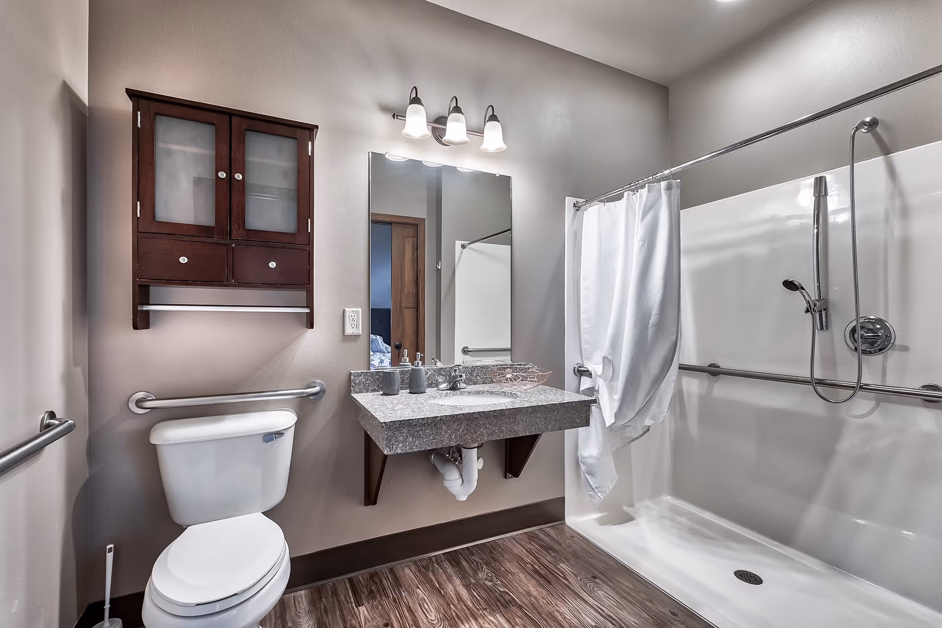 A clean and modern bathroom featuring a white toilet with grab bars on the wall, a granite countertop sink with a large mirror above it, a wooden cabinet mounted on the wall, and a walk-in shower with a white curtain and handheld showerhead. The floor has wood-like flooring and the walls are painted light gray.