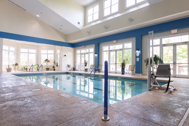 Bright indoor swimming pool area with a pool lift, seating, plants, and large windows.