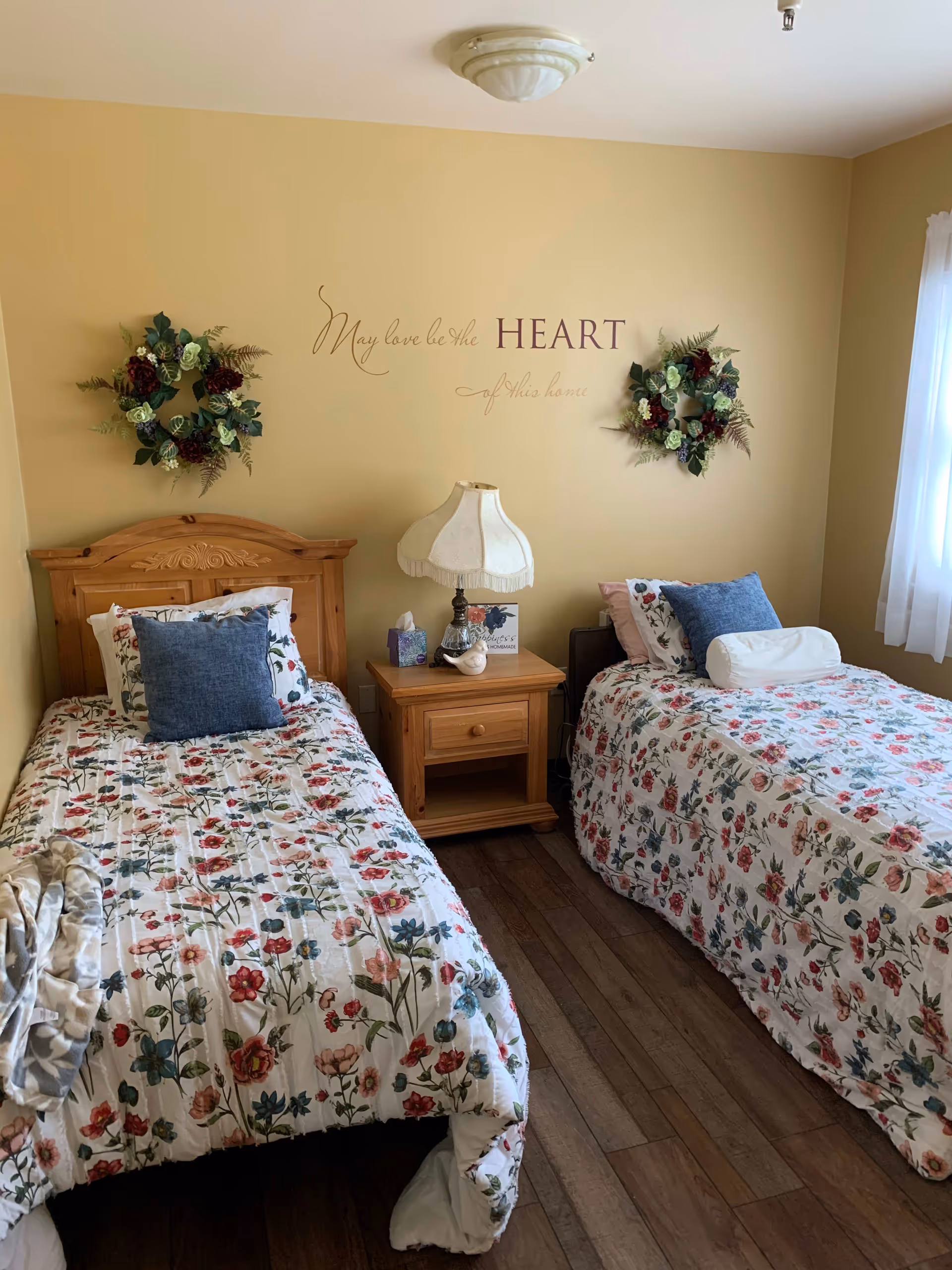 A cozy bedroom with two twin beds featuring floral bedspreads and blue accent pillows. A wooden nightstand with a vintage-style lamp, a tissue box, and decorative items sits between the beds. The walls are painted a warm beige color and decorated with two floral wreaths and a wall decal that reads 'May love be the HEART of this home.' A window with sheer white curtains allows natural light into the room.