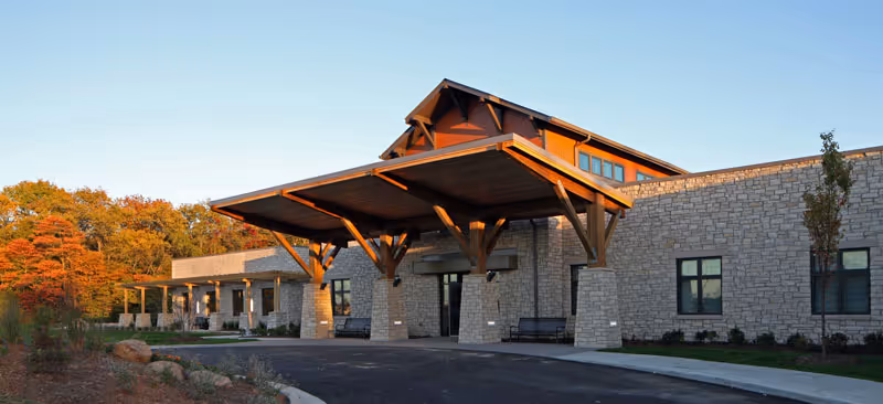 Exterior view of a senior living facility building with stone walls and a large wooden covered entrance. The building is surrounded by landscaped grounds and trees with autumn foliage under a clear sky.