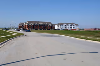 Wide paved road curving to the left with a large multi-story building complex in the distance under a clear blue sky. The building has red and white sections with multiple windows and a pitched roof. There is green grass on both sides of the road and a few parked vehicles near the building.