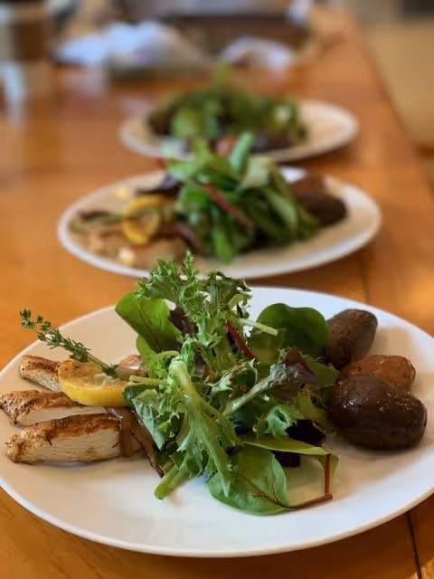 Close-up view of a white plate with grilled chicken breast, roasted baby potatoes, and a fresh green salad with mixed leafy greens and a lemon wedge, placed on a wooden table with two similar plates blurred in the background.