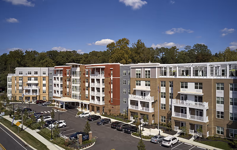 Exterior view of a modern multi-story senior living facility building with balconies, surrounded by trees and a parking lot with several cars under a blue sky with a few clouds.