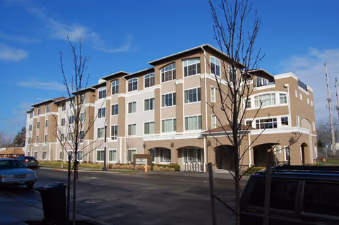 Exterior view of a multi-story senior living facility building under a blue sky with a few leafless trees in front and parked cars along the street.