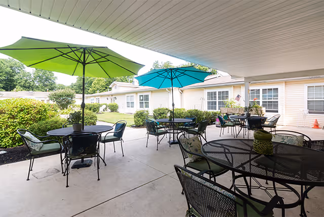 Covered courtyard patio with metal tables and chairs and green and teal umbrellas outside single-story residential buildings.