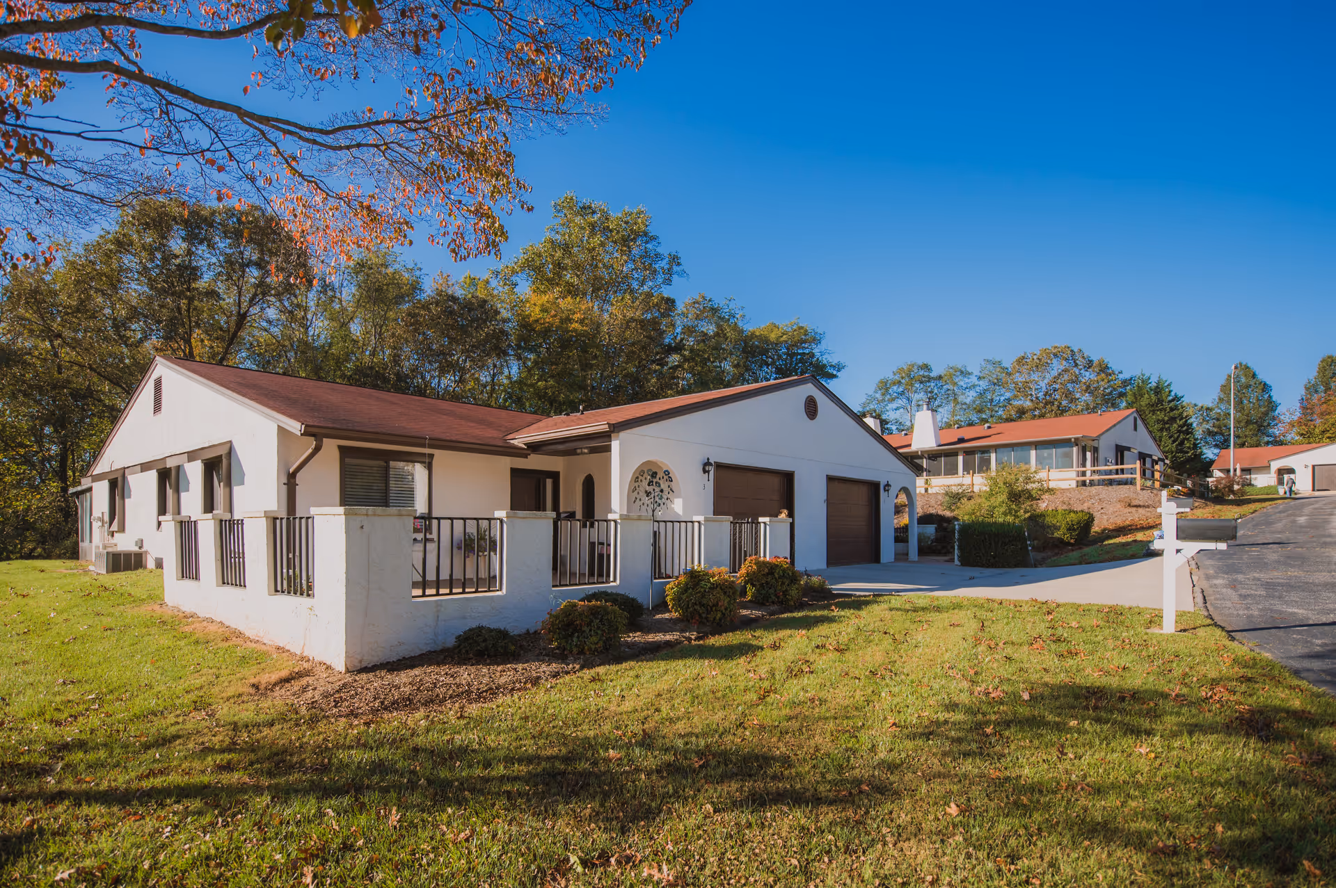 Exterior view of a single-story building with white walls and a red roof, surrounded by green grass and trees under a clear blue sky. The building has a small fenced patio area and a driveway leading to two garage doors. Additional similar buildings are visible in the background.