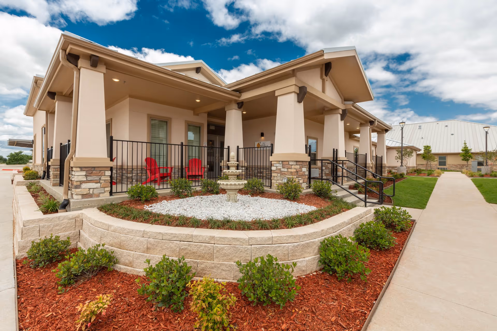 Exterior view of a single-story senior living facility building with beige walls, stone accents, and a covered porch area featuring red chairs. The landscaped garden in front includes a small fountain surrounded by white rocks and green shrubs, with a paved walkway leading to the entrance under a partly cloudy sky.