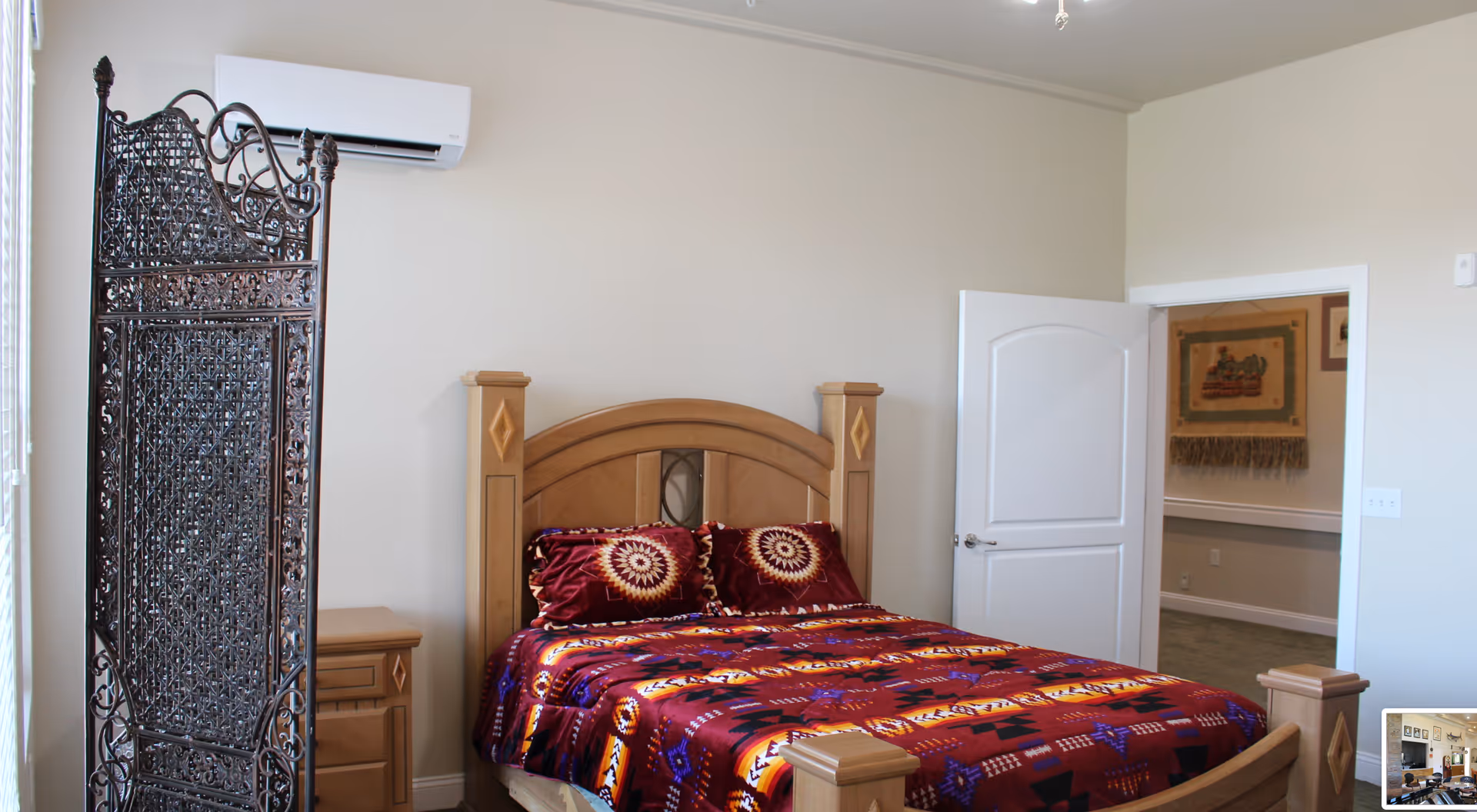A bedroom with a wooden bed frame and a colorful patterned bedspread in shades of red, orange, and purple. There are two matching pillows on the bed. To the left of the bed is a wooden nightstand and a decorative dark metal room divider. A white air conditioning unit is mounted on the wall above the bed. The room has beige walls and an open white door leading to another room with framed artwork on the wall.