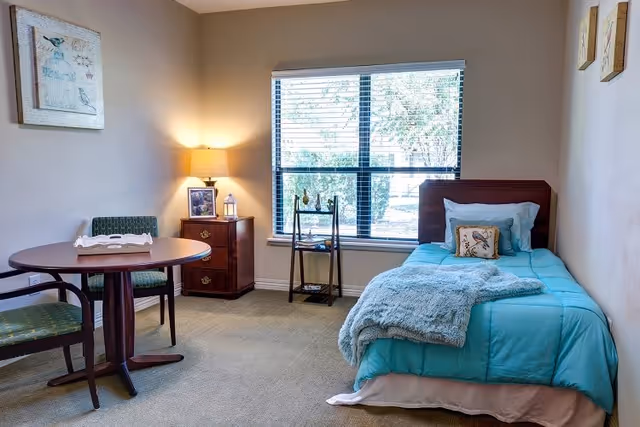 A cozy bedroom in a senior living facility featuring a single bed with turquoise bedding and a fluffy blanket, a small round table with two chairs, a wooden nightstand with a lamp and framed photo, a small shelving unit by the window, and wall art. The room has a large window with blinds letting in natural light.