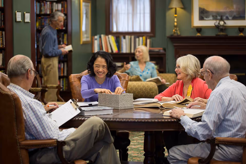 A group of older adults sitting around a table in a cozy common room, smiling and reading books.