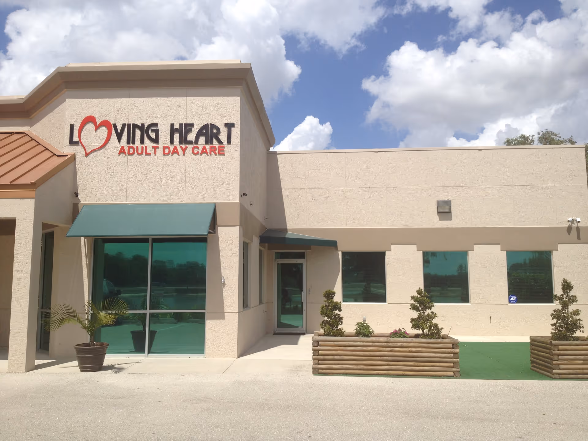 Exterior front view of Loving Heart Adult Day Care building with beige walls, green window awnings, potted plants, and a clear sky with clouds.