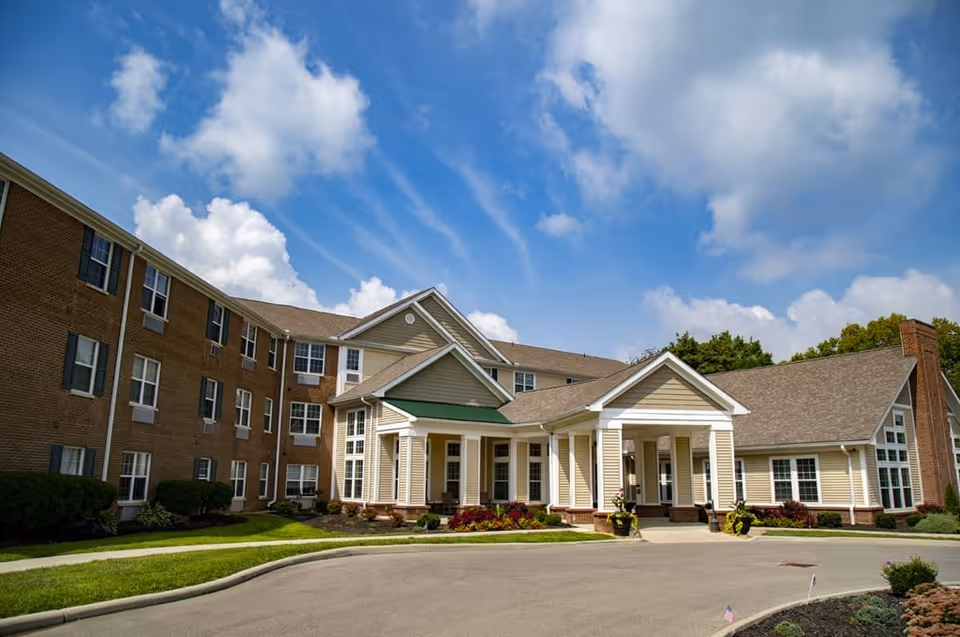 Exterior view of The Ashford of Mt. Washington senior living facility showing a large building with brick and beige siding, multiple windows, a covered entrance with white columns, landscaped greenery, and a partly cloudy blue sky.