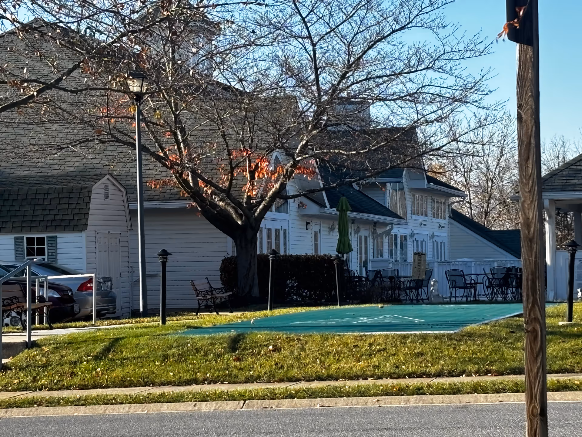 Outdoor area of a senior living facility with a shuffleboard court, several metal chairs and tables, a large tree with some remaining autumn leaves, and white buildings in the background under a clear blue sky.