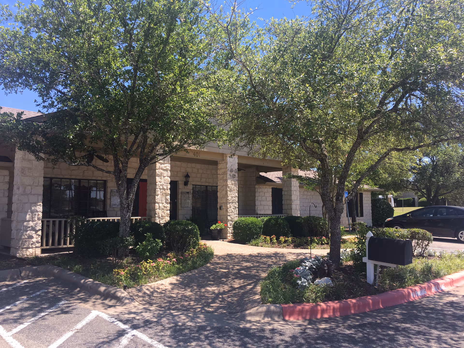 Exterior view of Wells Point Lodge Heatherwilde Assisted Living facility showing a stone building with a covered entrance, surrounded by trees, bushes, and a small garden area. A black mailbox is visible near the curb, and a parked car is seen in the background under a clear blue sky.