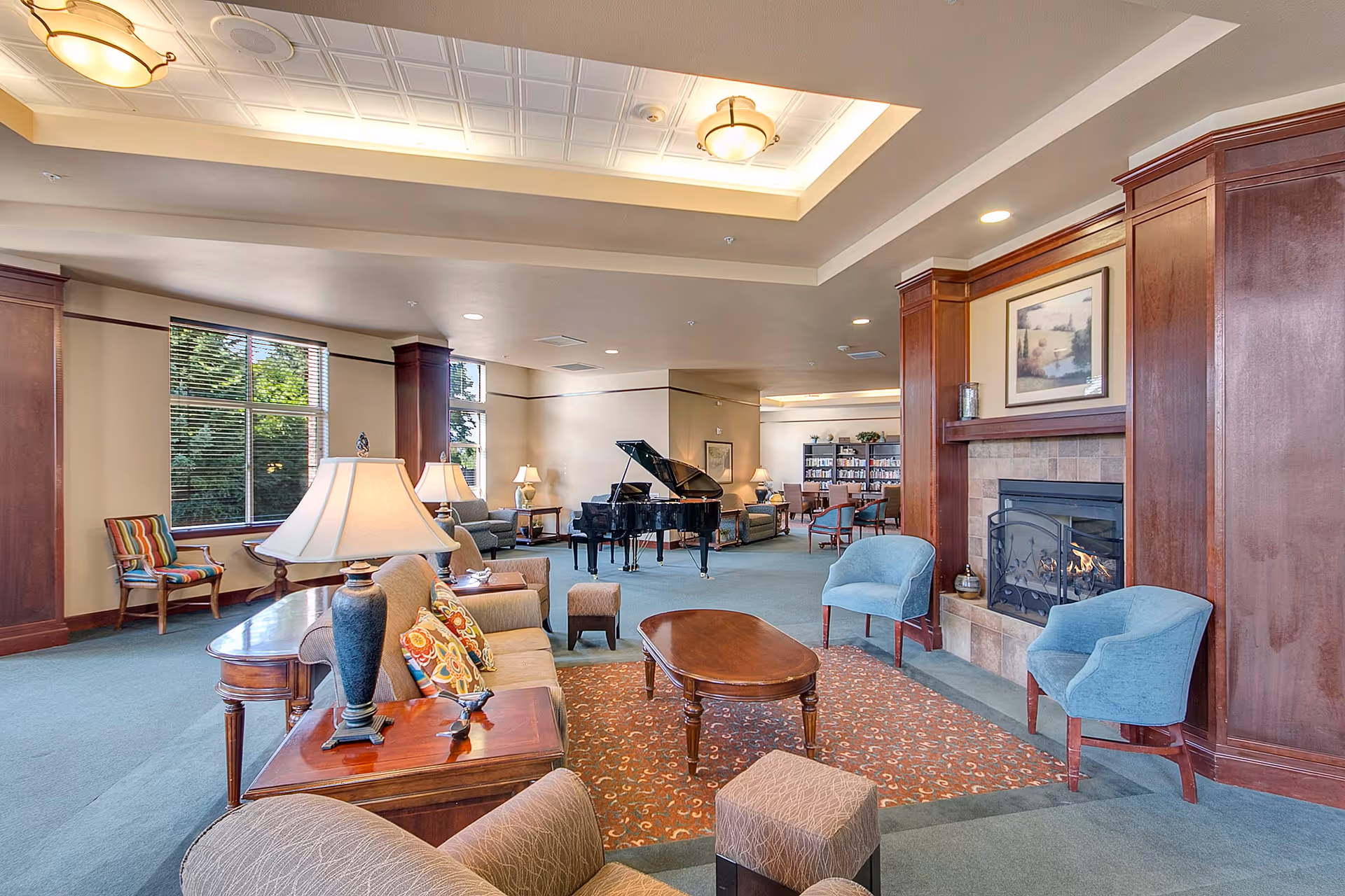 A spacious and well-lit living room area in a senior living facility featuring comfortable seating including sofas, armchairs, and ottomans arranged around a wooden coffee table and a fireplace with a framed picture above it. There is a grand piano in the background near large windows that let in natural light, and bookshelves with various items are visible further back in the room.